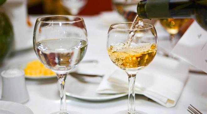 Sur une table un verre d'eau et un verre de vin blanc. Le verre de vin est en train d'être rempli.