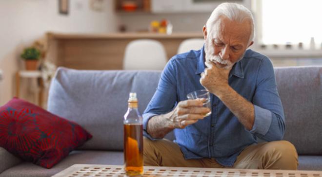 Un homme devant une bouteille de whisky et tenant un verre à la main. Il regarde son verre en se frottant le menton. Il semble dans une profonde réflexion.