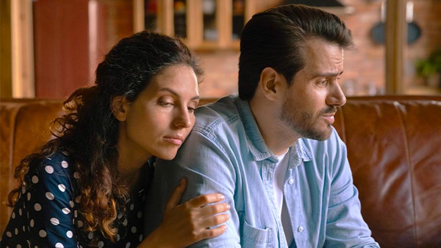 Un homme assis sur un canapé regarde dans le vague. Une femme assise à côté de lui le tient par l'épaule.